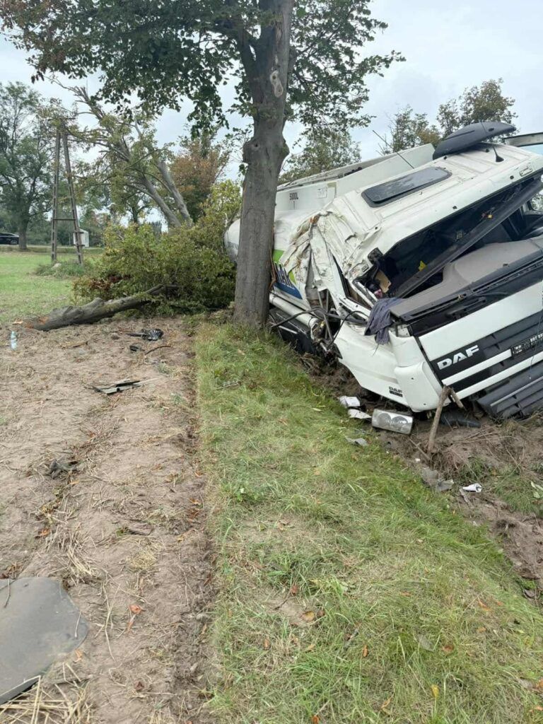 Złamany ciężarowy pojazd leży na boku obok drzewa, z uszkodzoną kabiną i porozrzucanymi elementami na trawie.