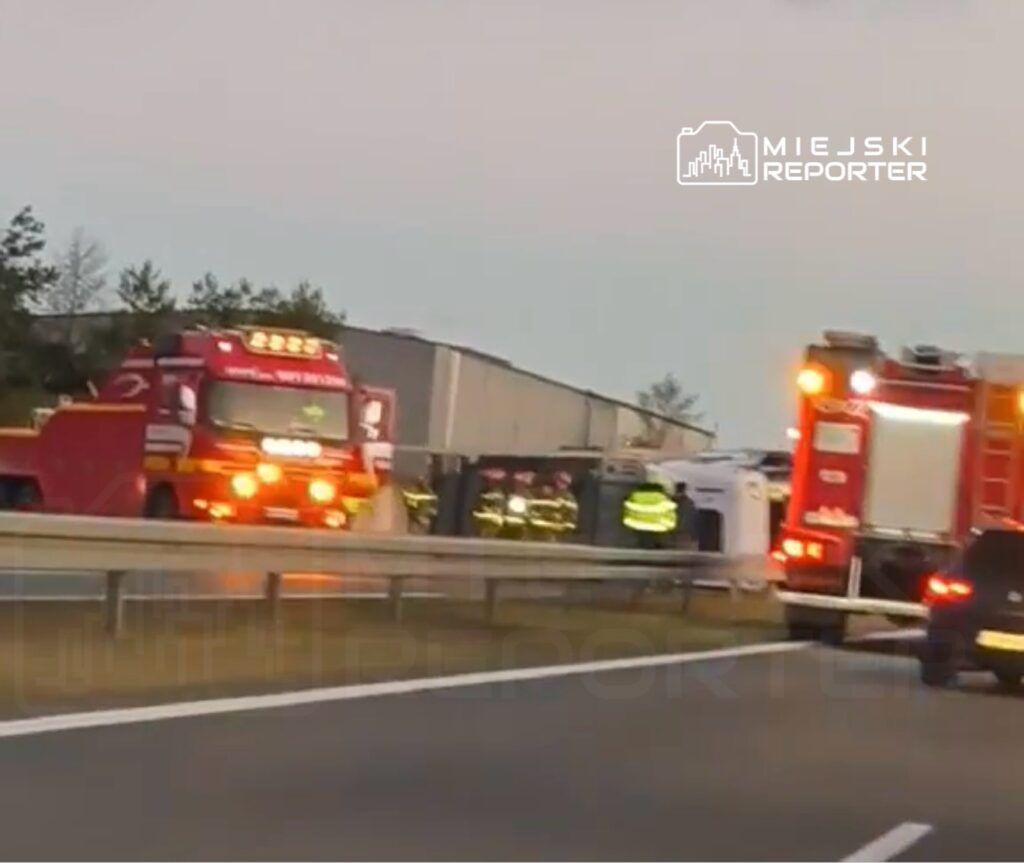 Czerwony wóz strażacki oraz zespół strażaków pracują przy przewróconym pojeździe na autostradzie.