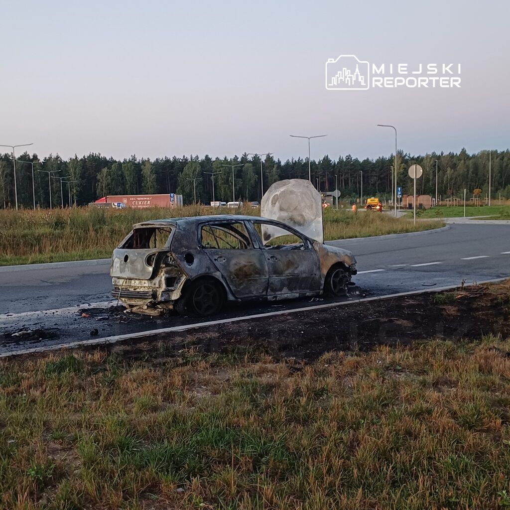 Spalony samochód osobowy zaparkowany na poboczu drogi, obok widoczna zieleń i znaki drogowe.