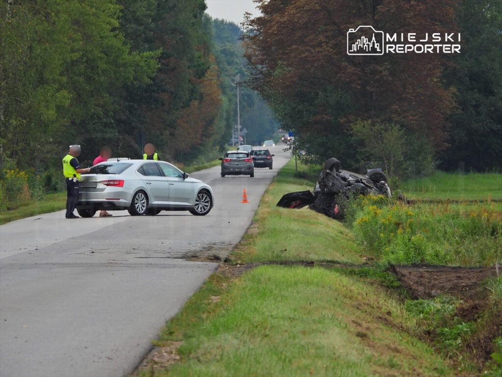 Policjant rozmawia z kierowcą srebrnego samochodu na drodze, obok przewrócone auto i znaki ostrzegawcze.