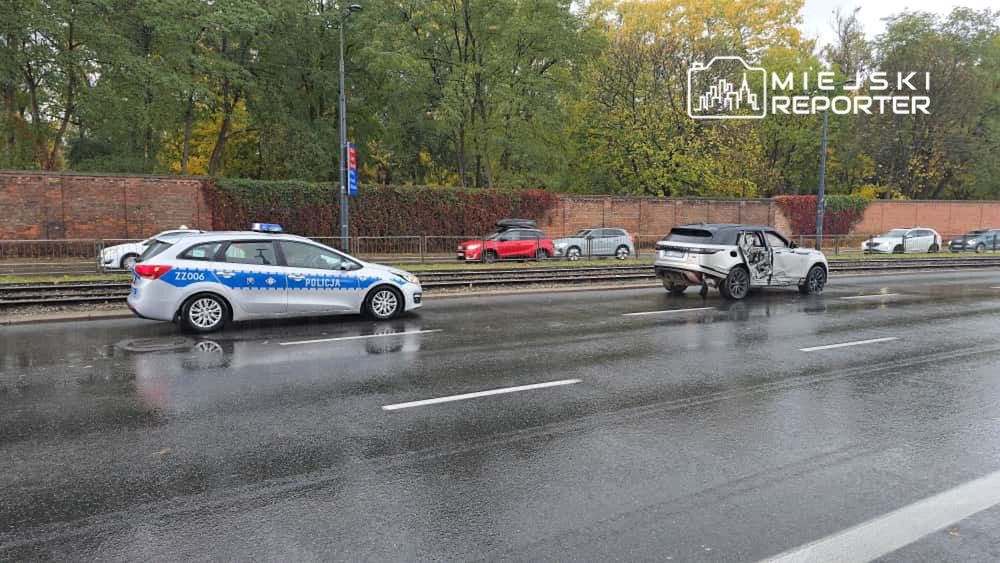 Policjant w radiowozie monitoruje sytuację na mokrej drodze, obok uszkodzony samochód z otwartymi drzwiami.