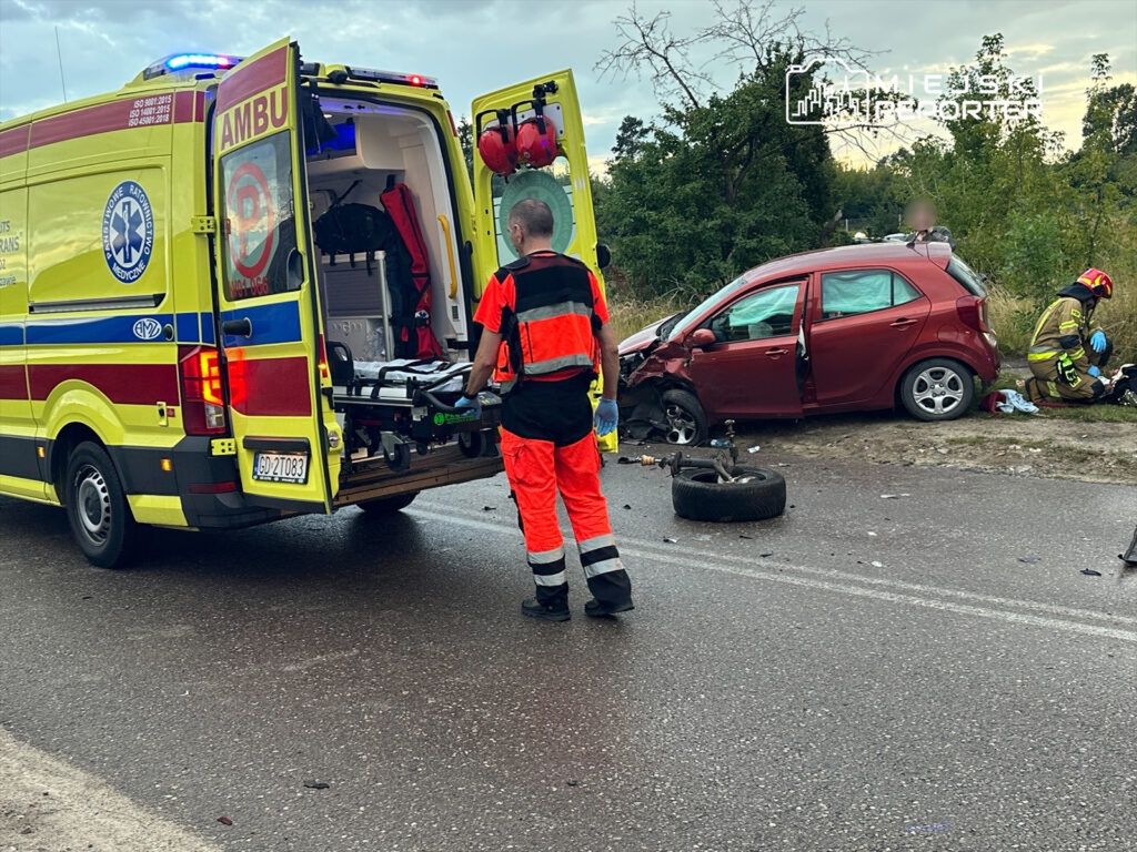 Ratownicy medyczni przygotowują nosze przy otwartej tylnej części karetki obok uszkodzonego czerwonego samochodu osobowego.