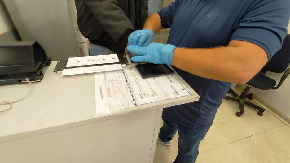 Mężczyzna w niebieskiej koszulce zakłada rękawiczki, przygotowując się do weryfikacji danych przy biurku.