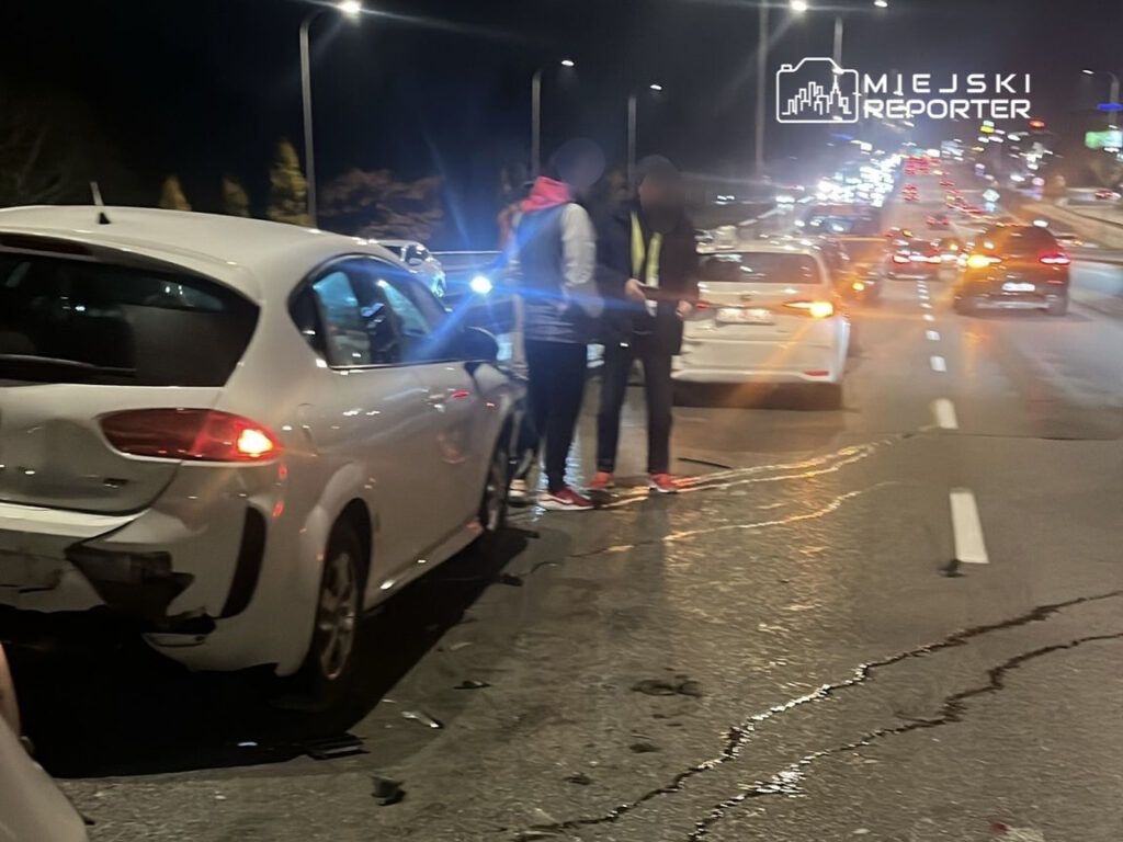 Dwaj mężczyźni rozmawiają obok uszkodzonego białego samochodu na ruchliwej drodze nocą.