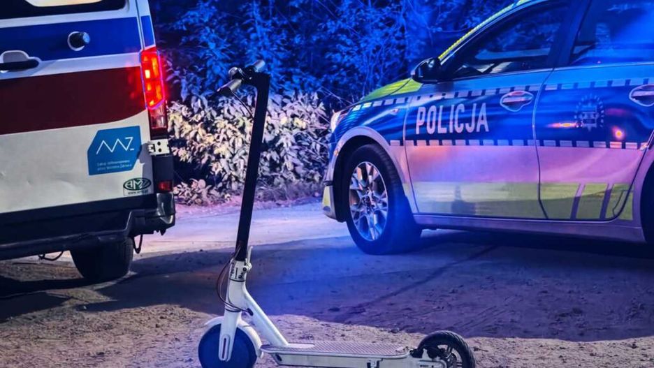 Skuter elektryczny stoi na drodze obok radiowozu policyjnego i karetki, otoczony niebieskim światłem.