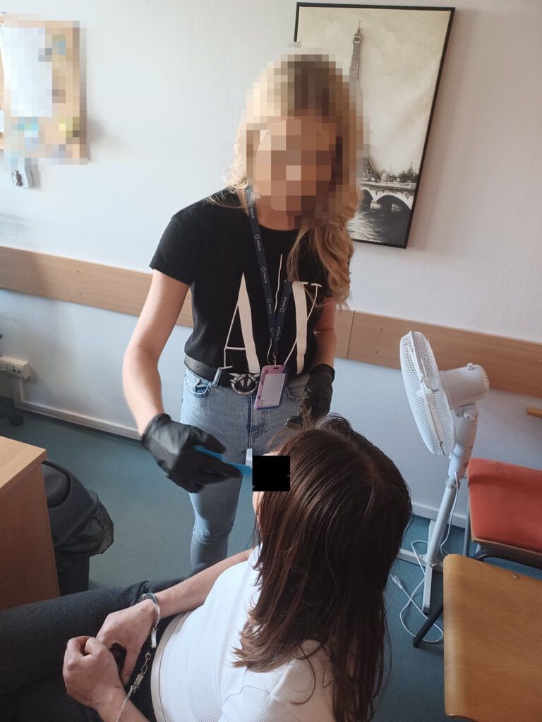 Kobieta w czarnej koszulce zakłada rękawice i wykonuje czynność na głowie siedzącej osoby w pomieszczeniu biurowym.