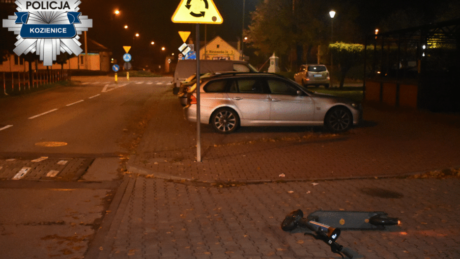 Na ulicy w nocy leży przewrócona hulajnoga elektryczna obok zaparkowanych samochodów. W tle widoczne znaki drogowe.