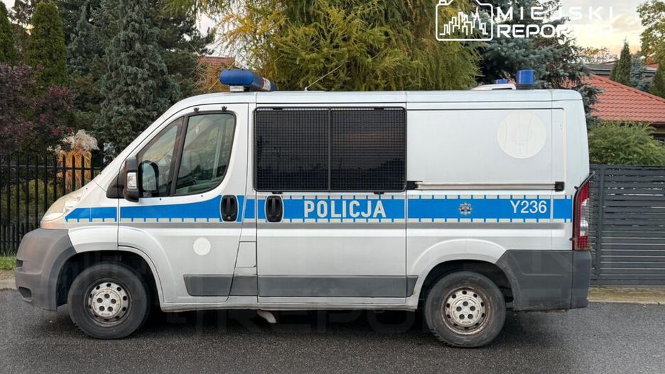 Funkcjonariusz policji w radiowozie parkowanym na ulicy, w tle ogrodzenie i drzewa.
