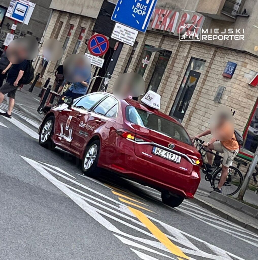 Czerwony samochód osobowy z oznaczeniem taksówki Uber stoi na skrzyżowaniu w mieście. W tle widoczni są piesi oraz rowerzysta, a także znaki drogowe informujące o przystanku autobusowym i zakazie parkowania. Scena przedstawia ruch uliczny w miejskim otoczeniu.