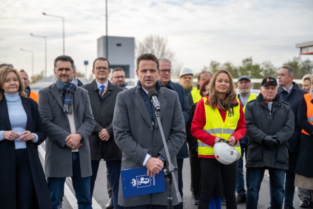 Mężczyzna w szarym płaszczu przemawia do zgromadzonego tłumu w kamizelkach odblaskowych na otwartej przestrzeni.