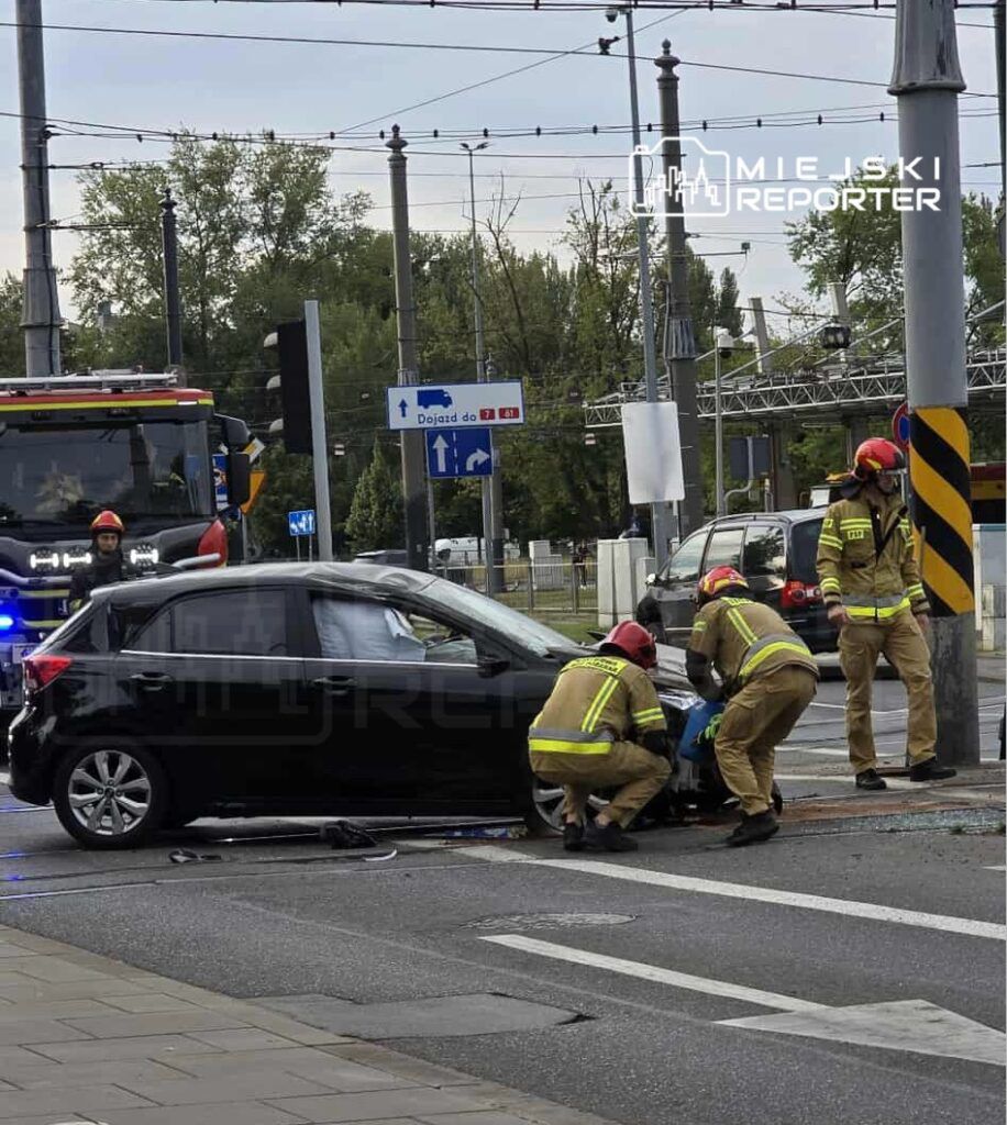 Czterech strażaków udziela pomocy poszkodowanemu przy czarnym samochodzie na ulicy, obok sygnalizacji świetlnej.