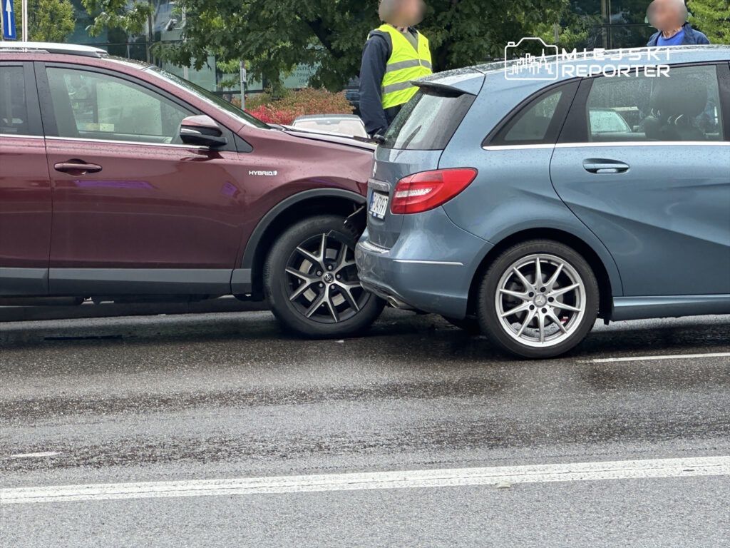 Dwa samochody osobowe zderzone na mokrej ulicy, obok stoi mężczyzna w żółtej kamizelce odblaskowej.