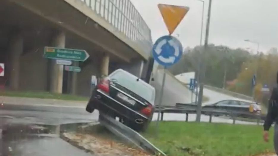 Czarny samochód osobowy unosi się na krawędzi drogi przy rondzie, obok znaku drogowym.