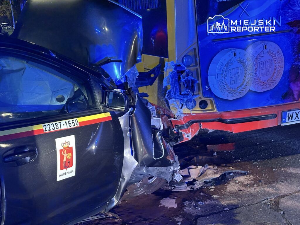 Policyjny samochód z uszkodzeniami tyłu zderza się z autobusem w nocy, oświetlonym niebieskimi światłami.