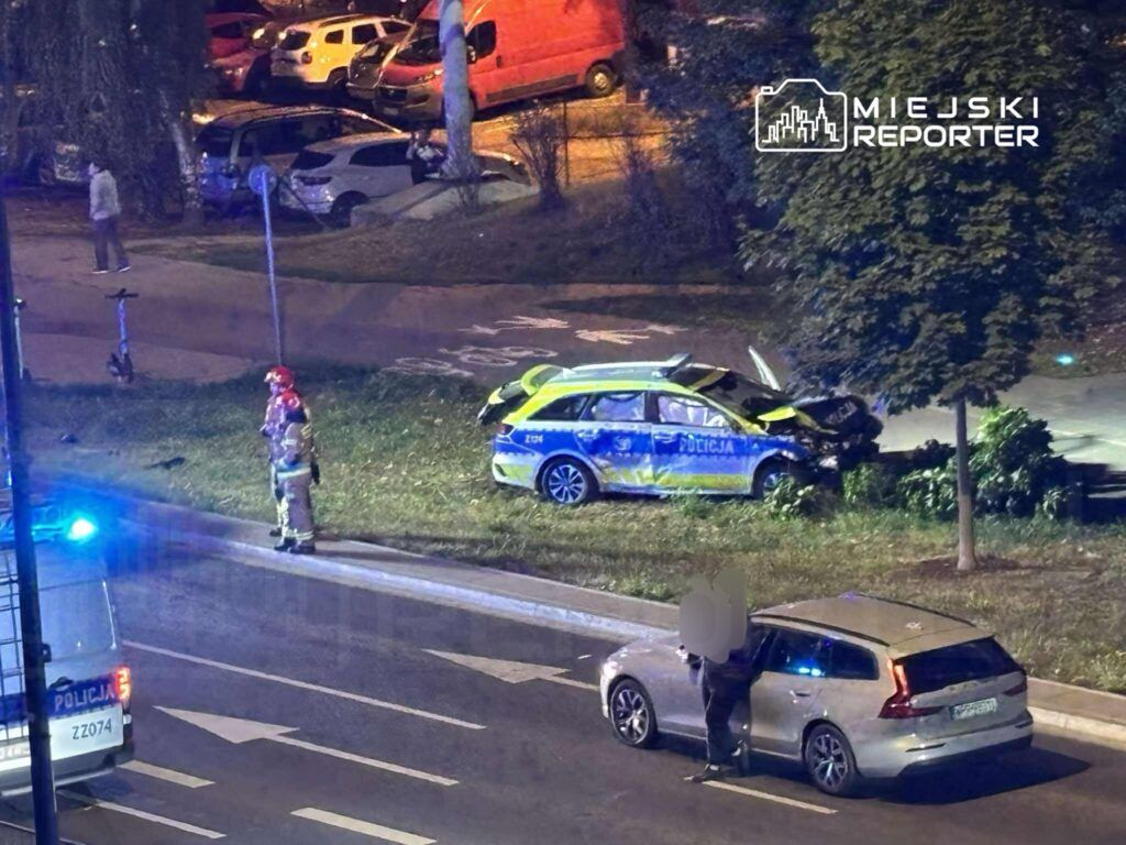 Policjant w mundurze stoi obok uszkodzonego radiowozu na trawie, a w tle widać zaparkowane samochody.