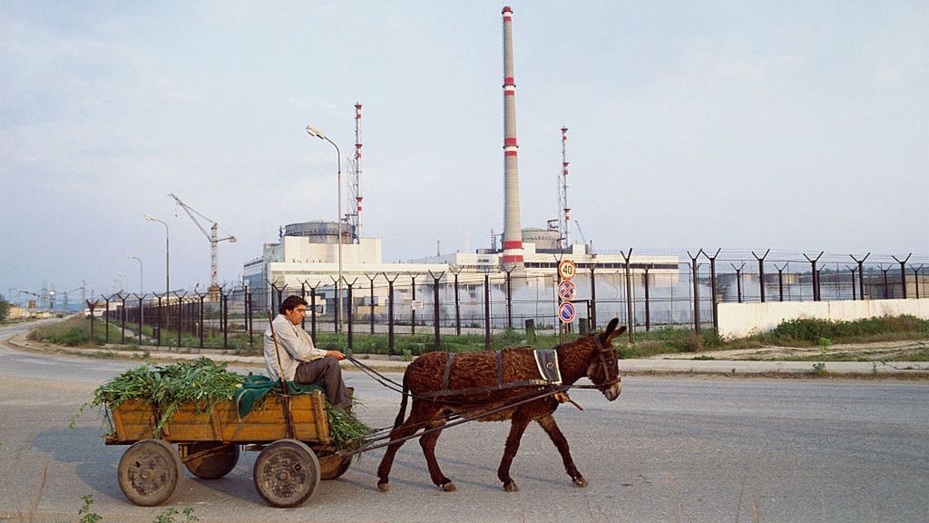 W tle elektrownia jądrowa w Kozłoduju. Na pierwszym planie rolnik na ciągniku, w który zaprzągnięty jest osioł