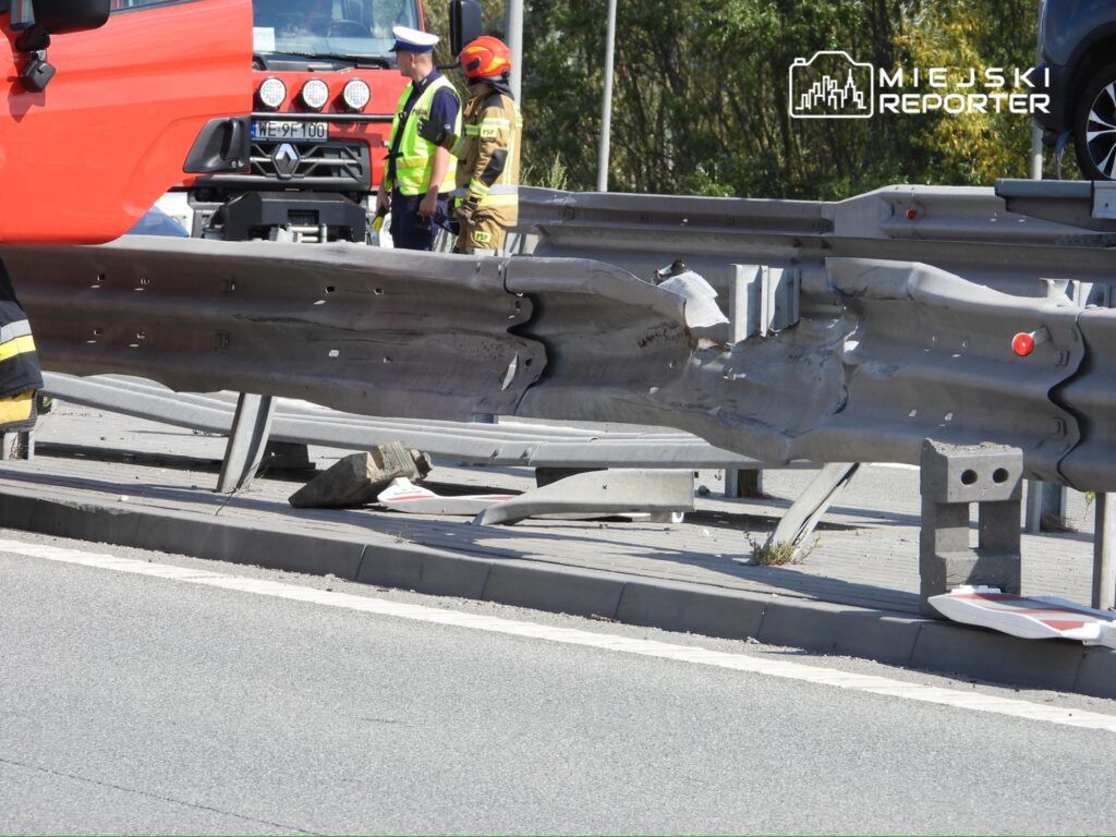 Uszkodzony barierka ochronna na drodze, w pobliżu pracujących strażaków i policjantów, z fragmentami gruzu na jezdni.