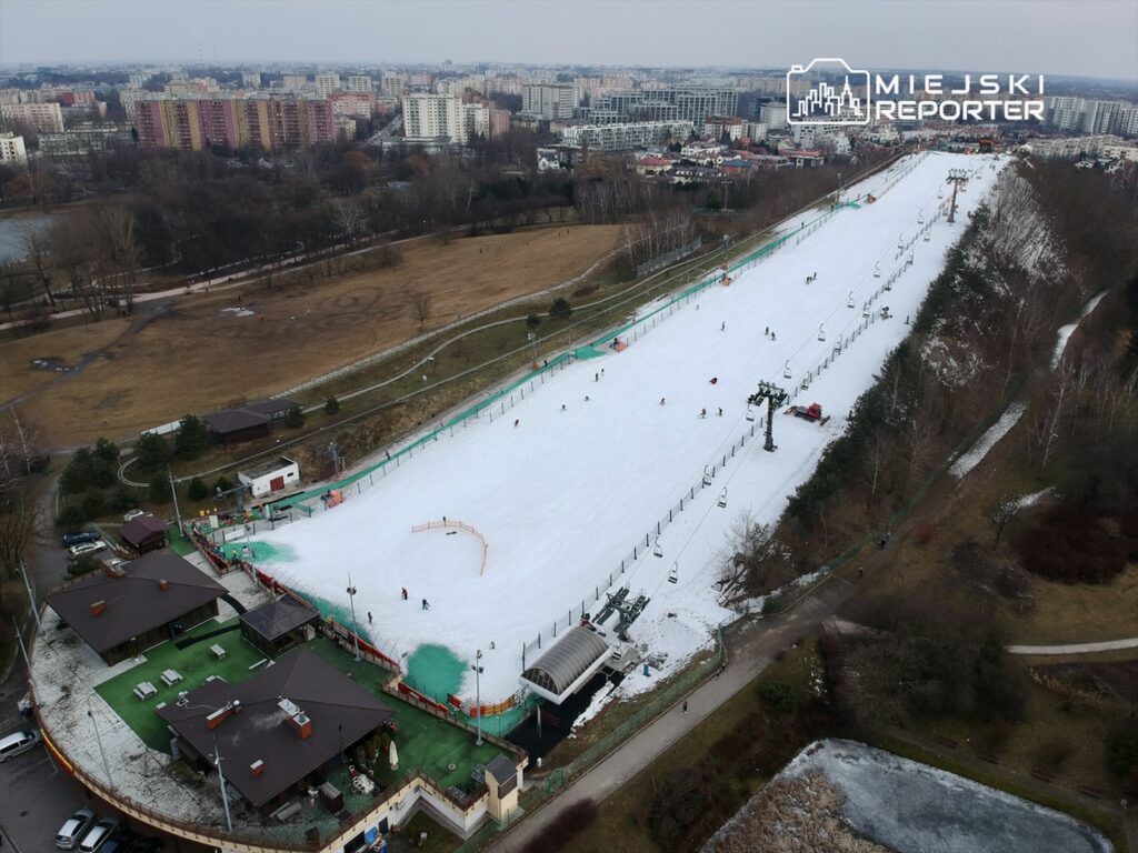 Narciarze zjeżdżają po stoku w mieście, obok nowoczesnych budynków i terenów zielonych. W tle widoczny wyciąg narciarski.