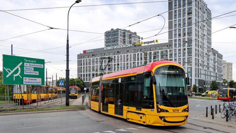 Tramwaj w kolorze żółto-czerwonym skręca na skrzyżowaniu, a w tle widać nowoczesne budynki biurowe.
