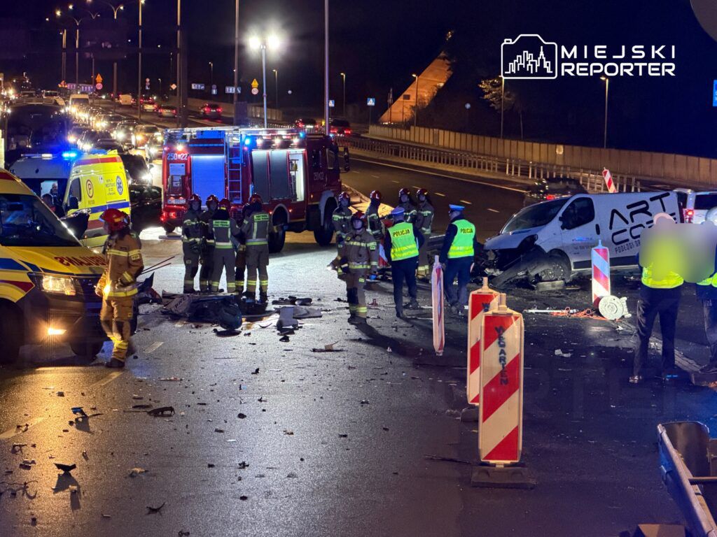 Grupa ratowników i policjantów pracuje na miejscu wypadku drogowego, obok uszkodzonego furgonu na ruchliwej drodze nocą.