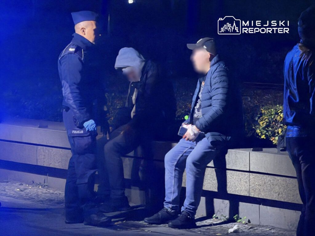Policjant w mundurze rozmawia z dwoma mężczyznami siedzącymi na ławce w ciemnej okolicy, oświetlonej niebieskim światłem.