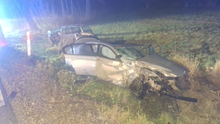 Uszkodzony samochód osobowy leży w rowie obok drogi, z widocznymi uszkodzeniami karoserii. W tle widać drugi pojazd.