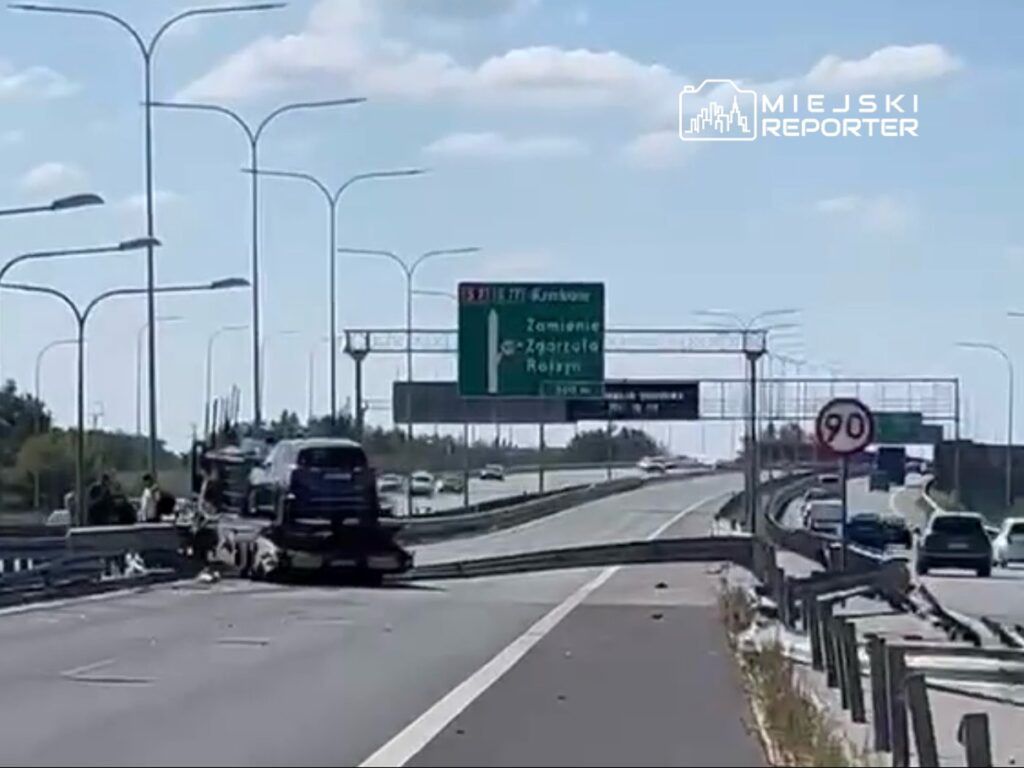 Na autostradzie stoi uszkodzony samochód ciężarowy, a obok grupka osób rozmawia. W tle widoczne znaki drogowe.