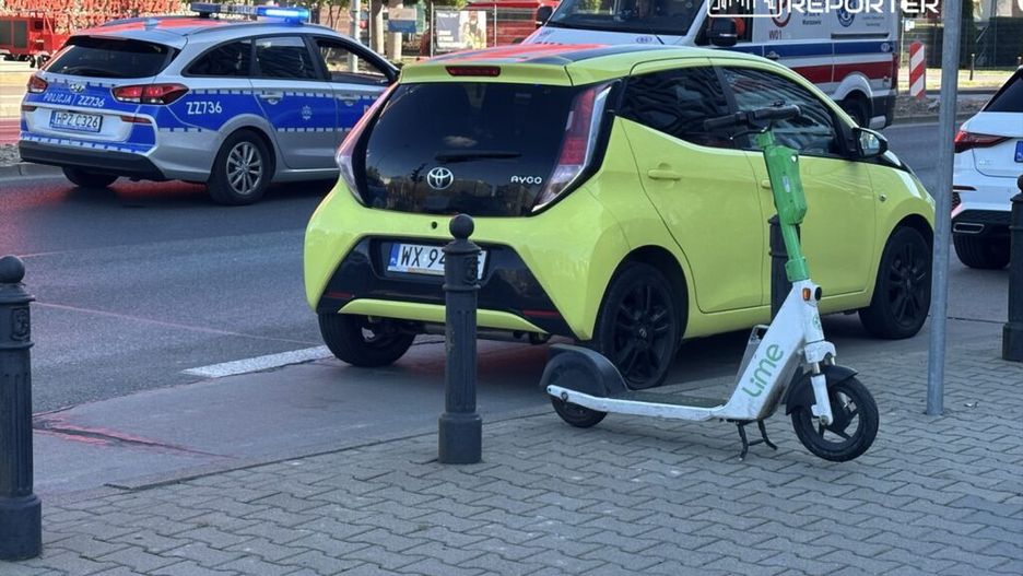 Żółty samochód osobowy zaparkowany obok skutera elektrycznego Lime, w tle radiowóz policyjny z włączonymi sygnałami świetlnymi.