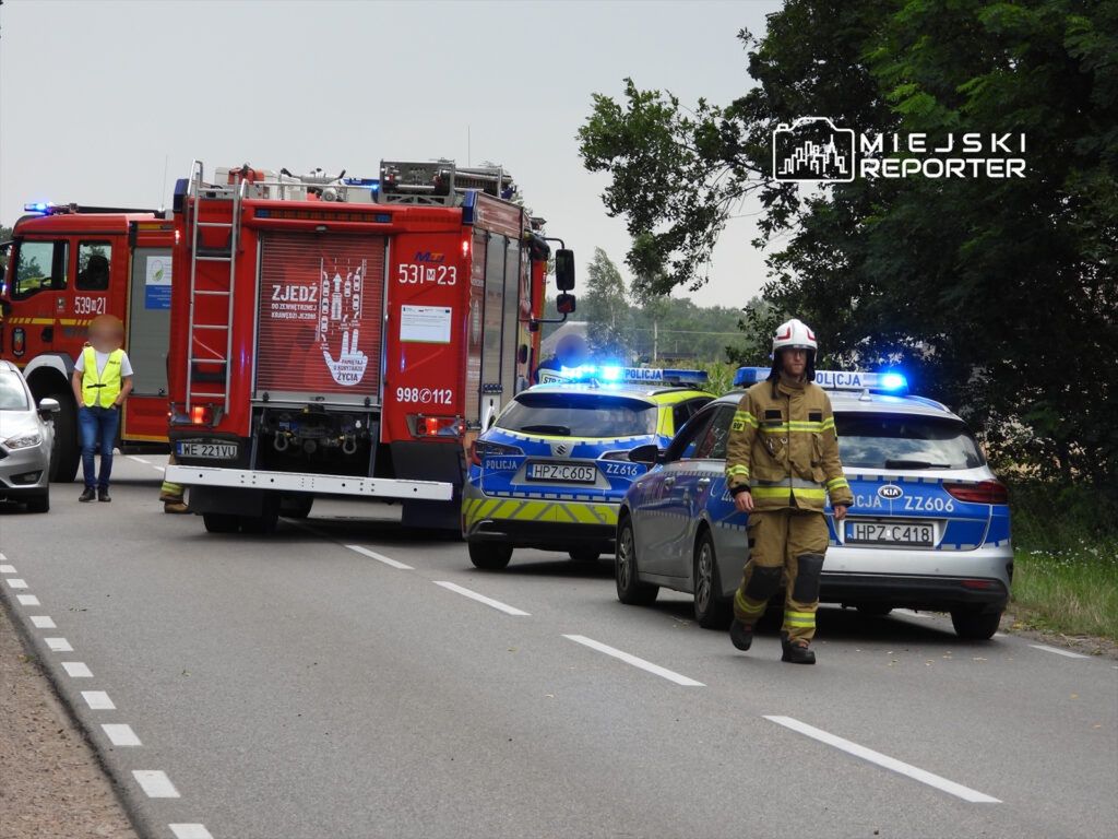 Strażak w mundurze kieruje ruchem obok wozu strażackiego i radiowozów policyjnych na drodze.