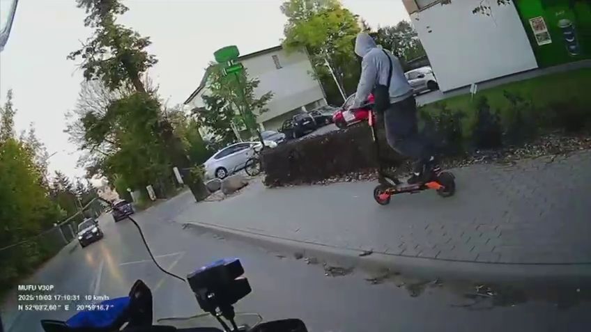 Mężczyzna w szarej bluzie jedzie na hulajnodze elektrycznej po chodniku w miejskim otoczeniu.