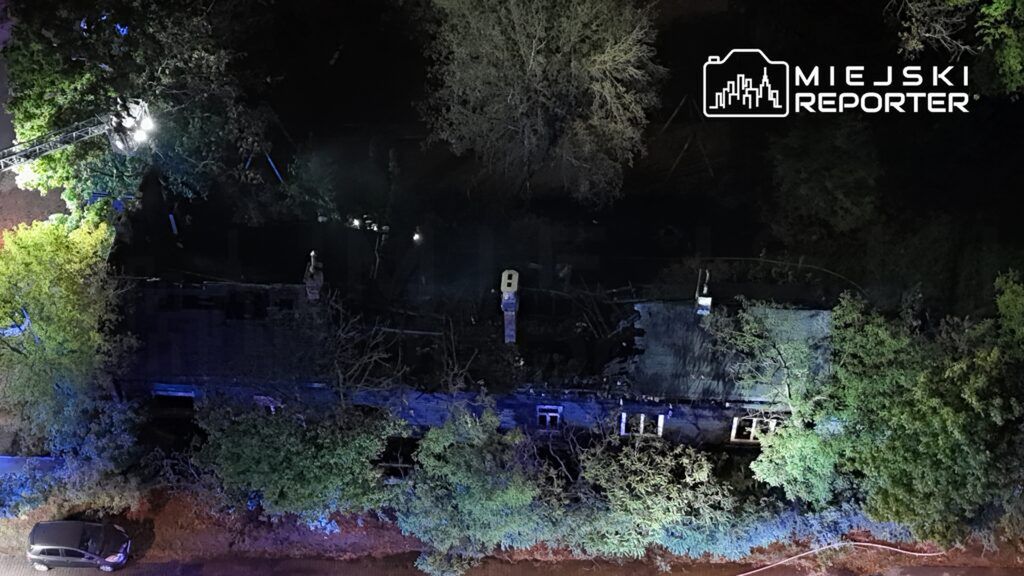 Zniszczony budynek otoczony przez drzewa, oświetlony niebieskimi i białymi światłami, z widocznymi zniszczeniami na dachu.