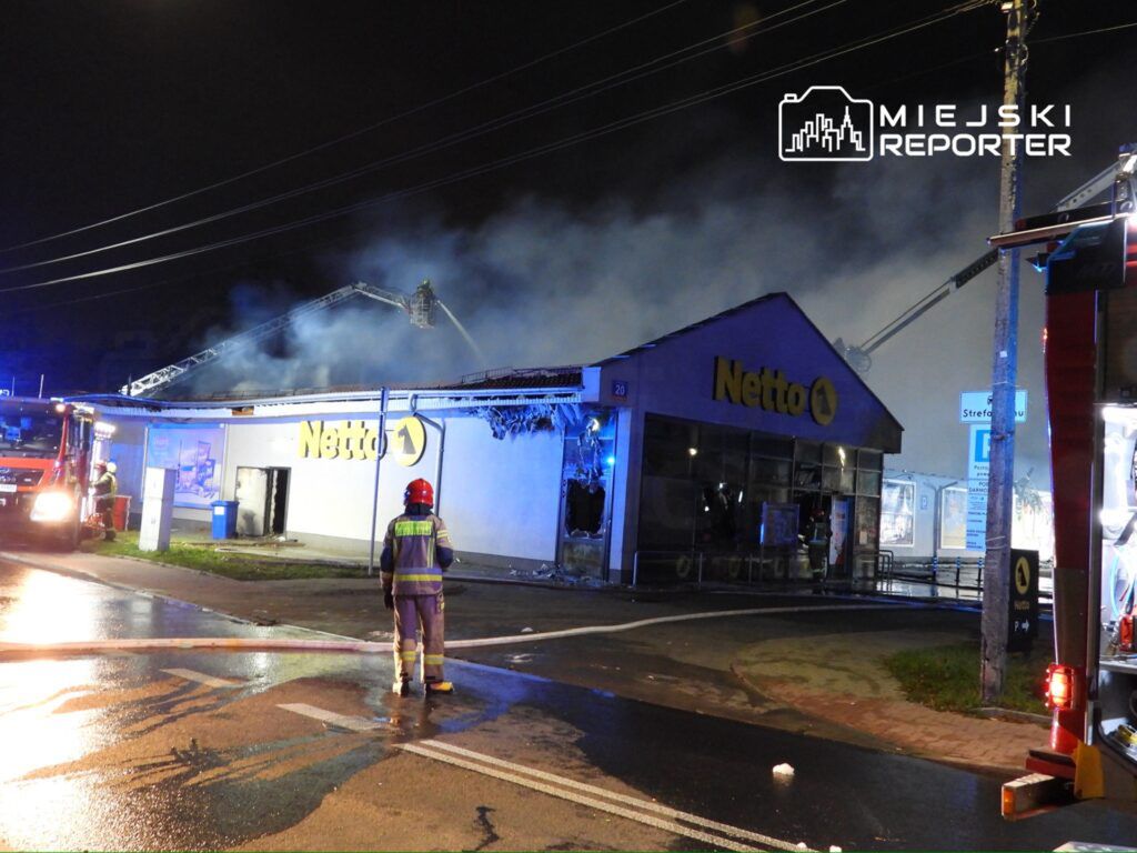 Strażacy gaszą pożar w sklepie Netto, z którego wydobywa się dym, w nocy przy oświetlonych ulicach.