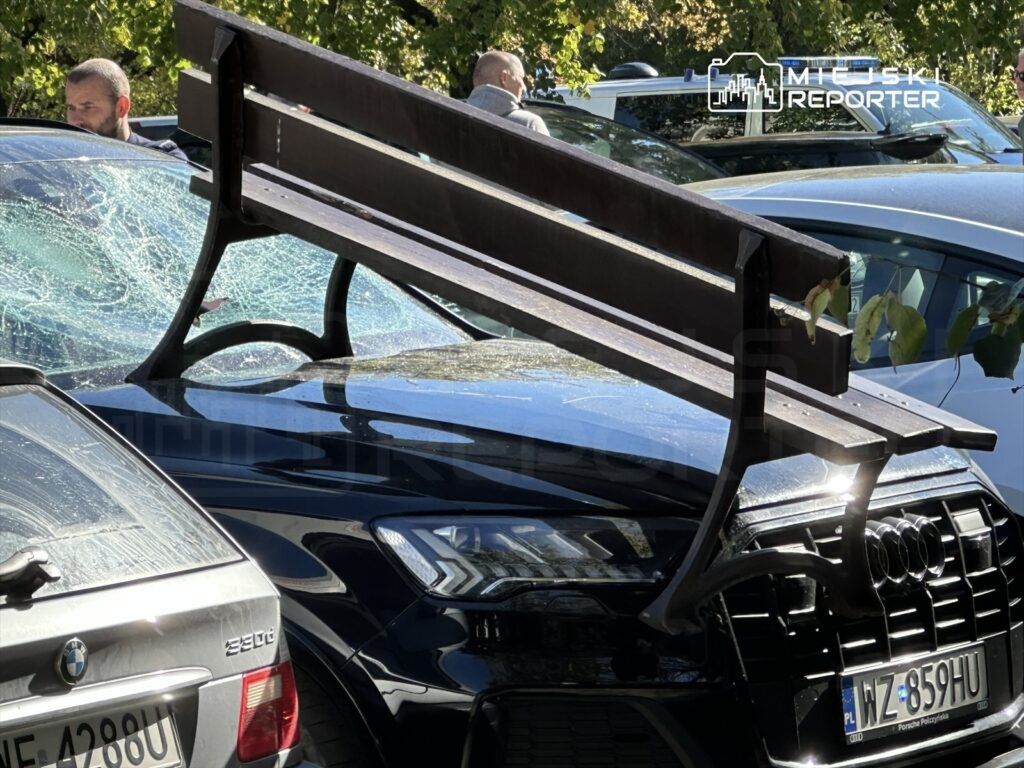 Złamana ławka leży na dachu czarnego samochodu, obok stoją mężczyźni, a w tle widać inne pojazdy.