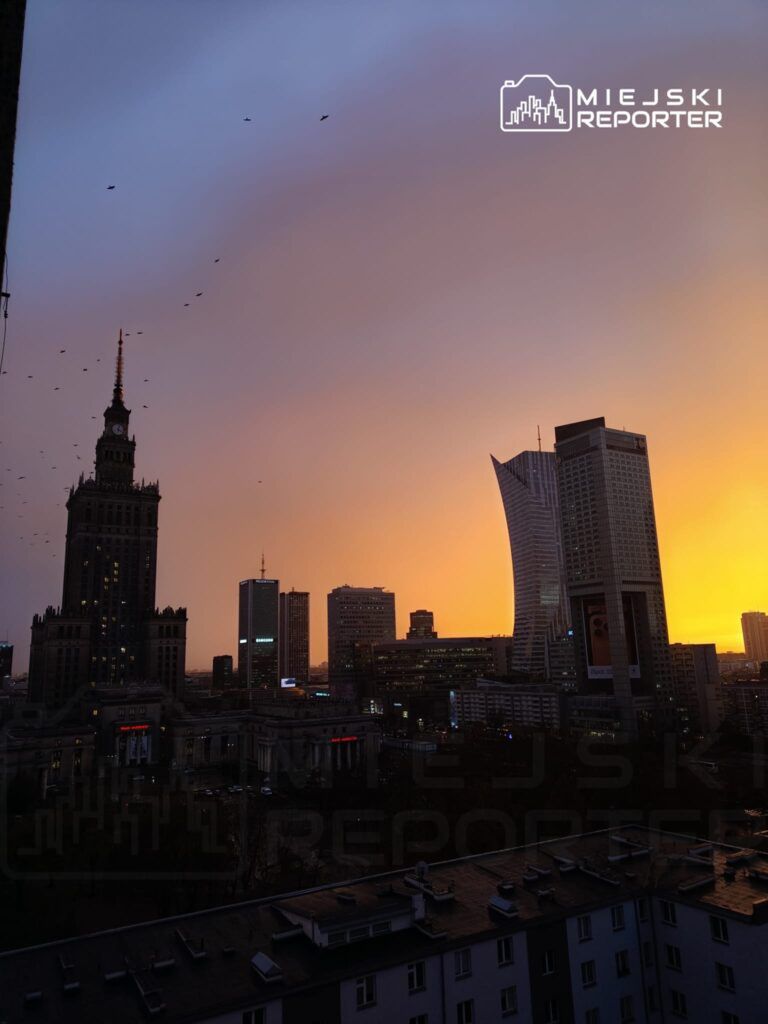 Zachód słońca nad miastem, z widokiem na wysokie budynki i charakterystyczną wieżę w tle.