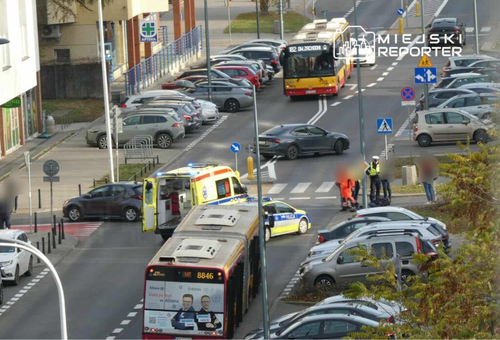 Na skrzyżowaniu zatrzymane pojazdy, w tym karetka i radiowóz, obok grupka osób w odblaskowych kamizelkach.
