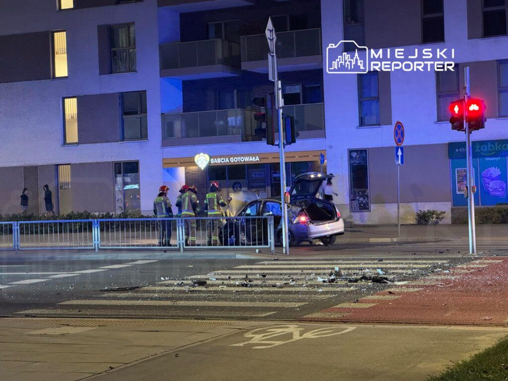 Grupa strażaków w hełmach pracuje przy uszkodzonym samochodzie na skrzyżowaniu, obok sklepu spożywczego.