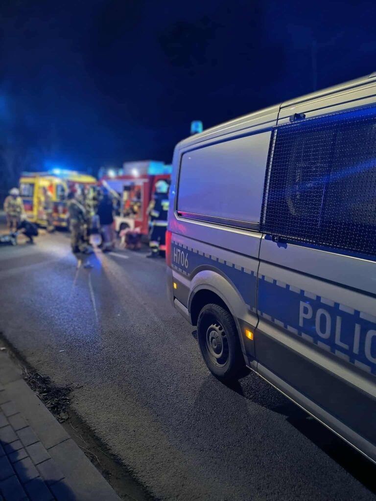 Na drodze nocą interweniują policjanci oraz strażacy, obok zaparkowany radiowóz i woz strażacki.