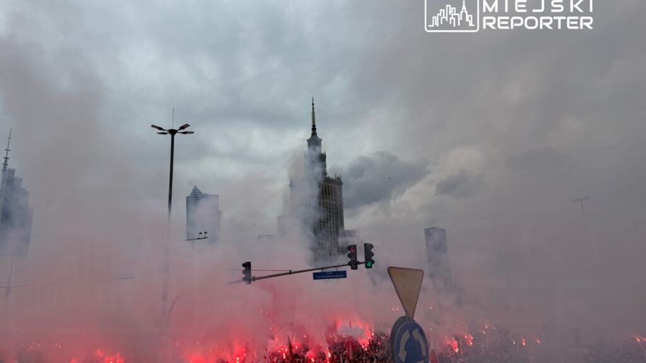 Tłum ludzi z flagami w Polsce, otoczony dymem z rac, na ulicy w mieście, w tle wieżowiec.