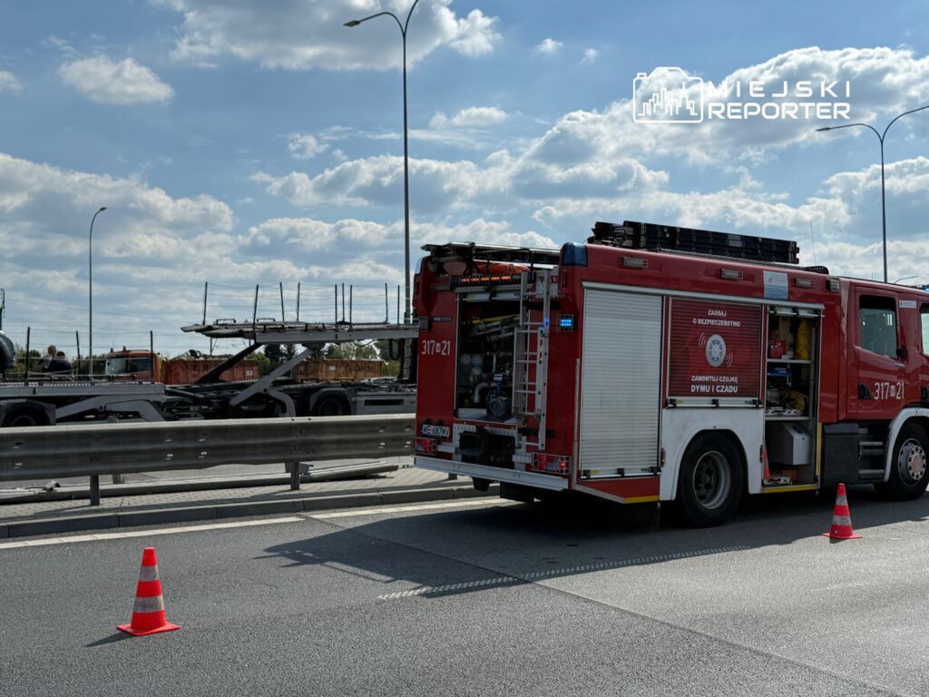 Wóz strażacki z otwartymi drzwiami, zaparkowany na pasie drogi, obok trójkąt ostrzegawczy. W tle widoczny transport ciężarowy.