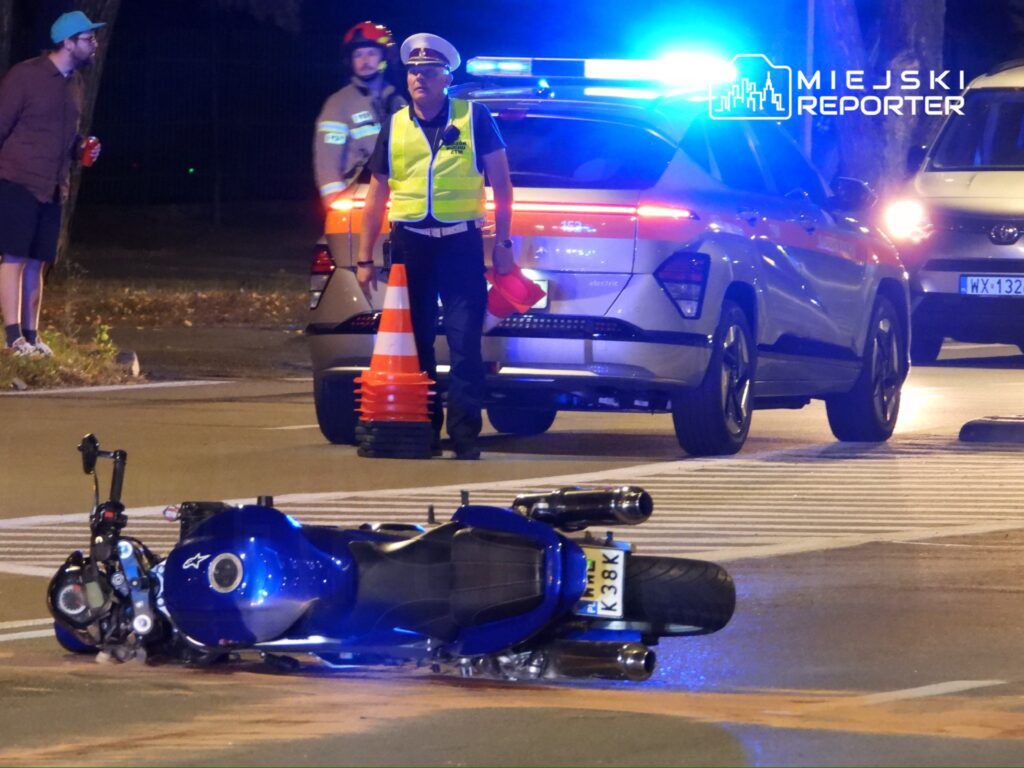 Motocykl leżący na boku na drodze, obok stoi policjant w kamizelce odblaskowej, a w tle widać radiowóz.