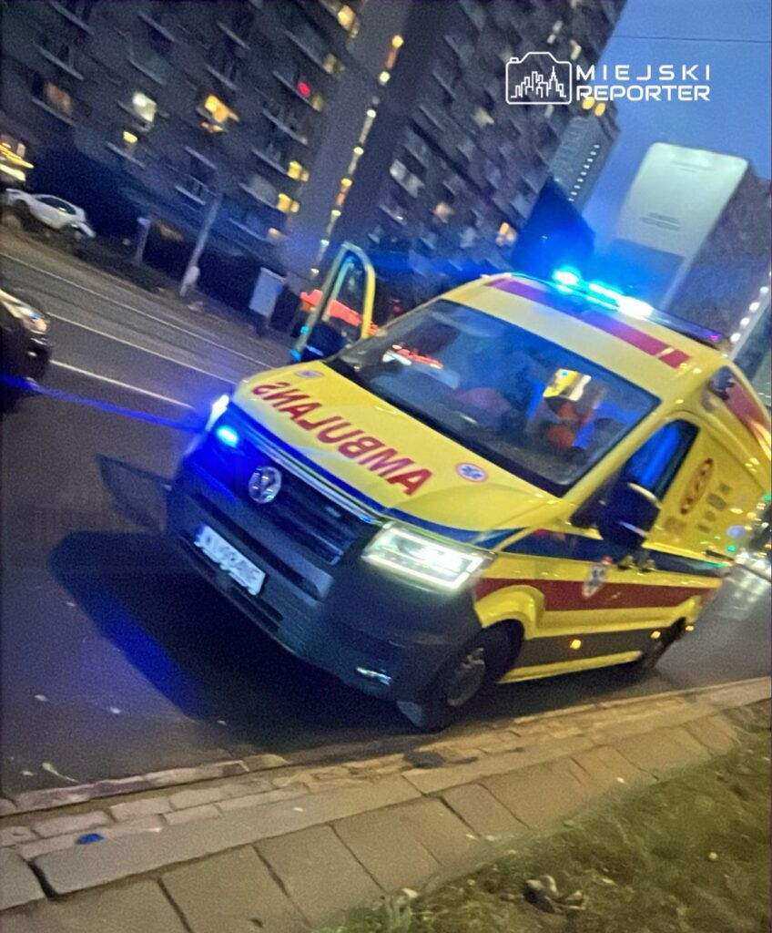 Karetka pogotowia z włączonymi sygnałami świetlnymi zatrzymana na ulicy w pobliżu wysokiego budynku mieszkalnego.