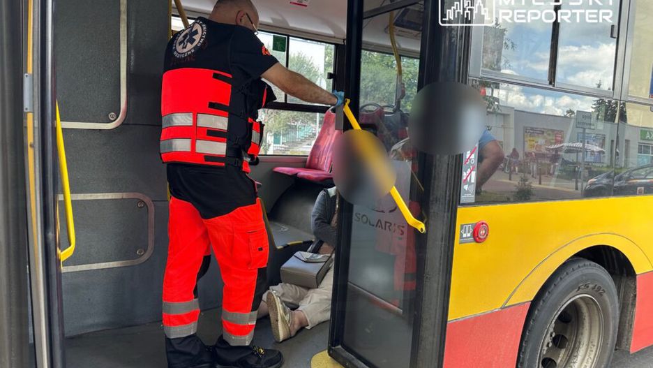 Ratownik medyczny w odblaskowej kamizelce udziela pomocy osobie siedzącej na podłodze autobusu miejskiego.
