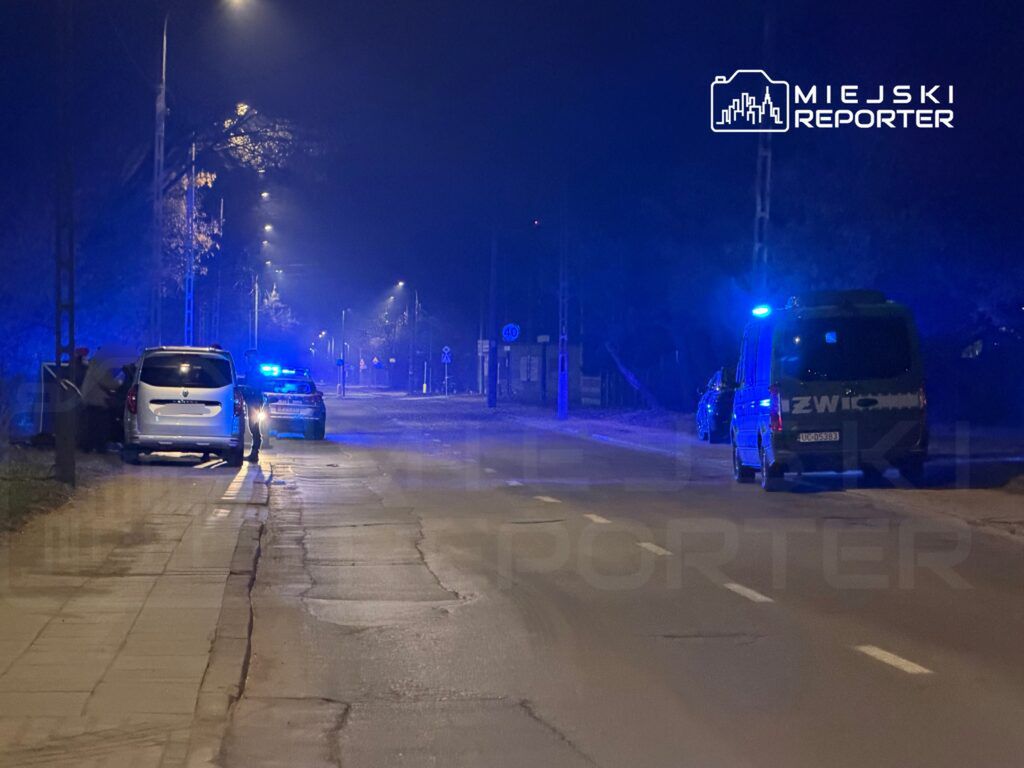 Nocna ulica oświetlona niebieskimi światłami radiowozów policji i pojazdu specjalistycznego, zaparkowane samochody.