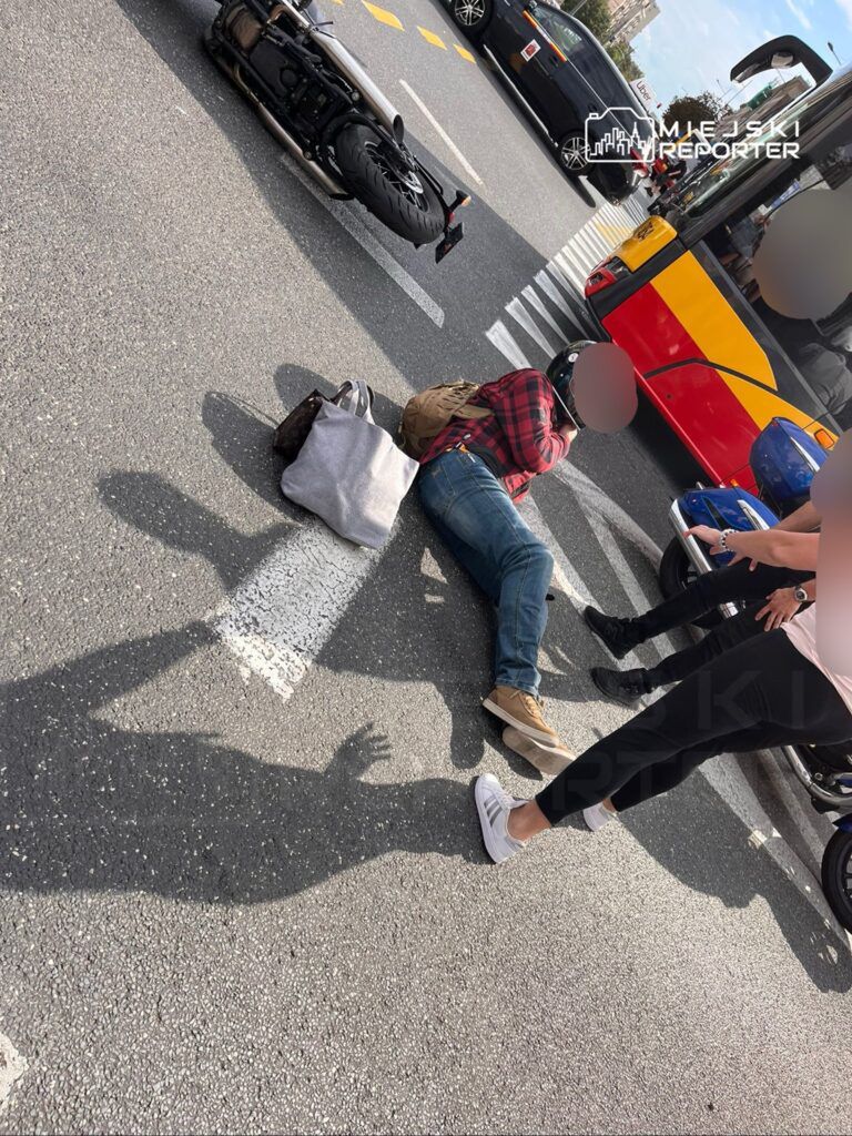 Wypadek drogowy z udziałem motocykla i pieszych na ulicy. Motocykl leży na jezdni, a poszkodowany mężczyzna znajduje się obok, trzymając się za głowę. W tle widać autobusy i inne pojazdy. Obecni świadkowie próbują udzielić pomocy. Sytuacja wywołuje zaniepokojenie przechodniów.