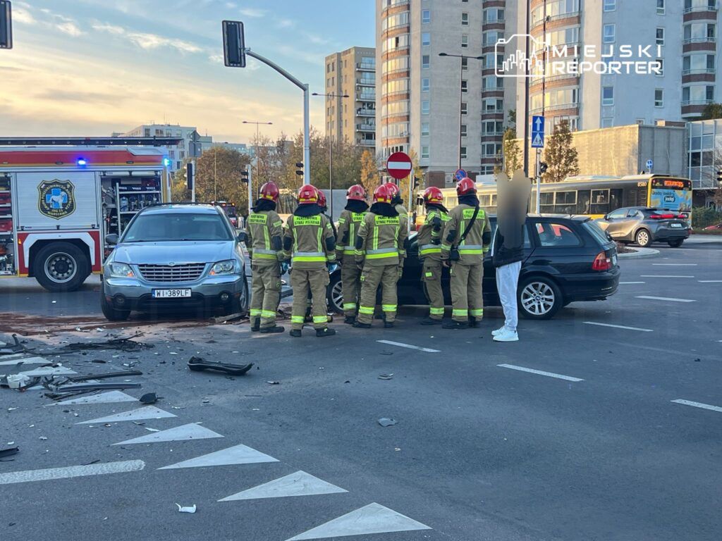 Grupa strażaków w kaskach omawia sytuację przy zderzeniu dwóch samochodów na skrzyżowaniu, obok wozu strażackiego.