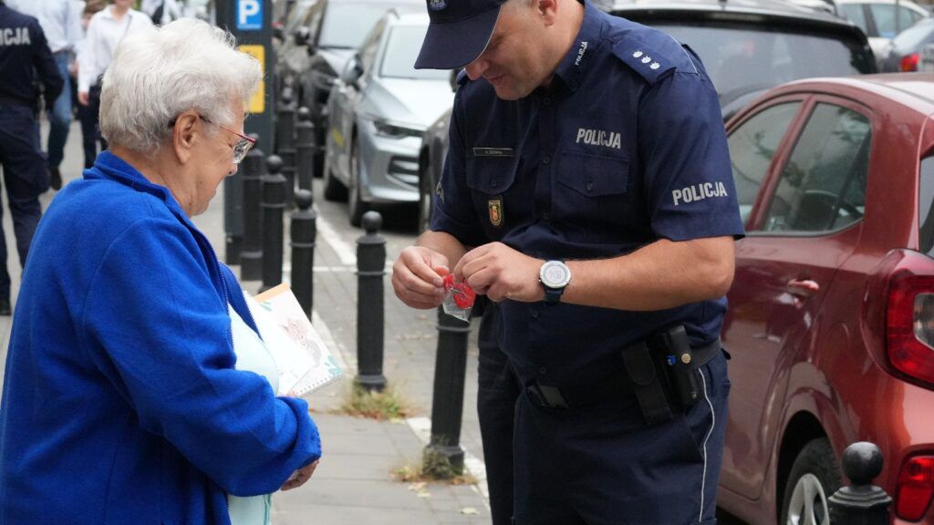 Starsza kobieta w niebieskiej kurtce odbiera od policjanta czerwoną zawieszkę na ulicy, w tle samochody.