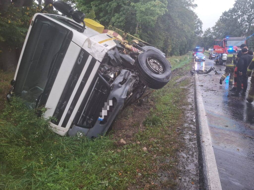 Cysterna przewrócona na bok na poboczu drogi, obok strażacy i pojazdy ratunkowe w deszczowej aurze.