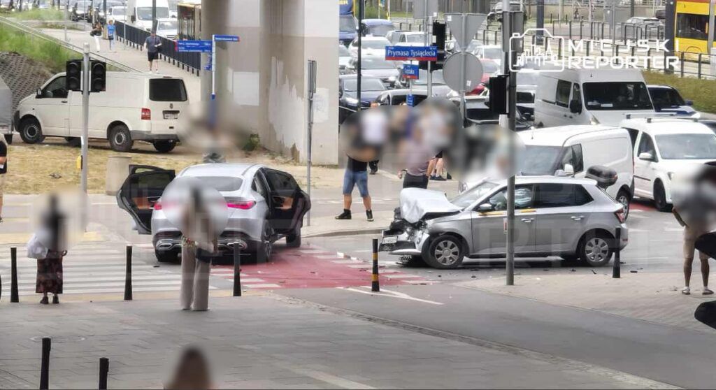 Kobieta z torbą stoi na przejściu dla pieszych, obok zderzone samochody, a w tle ruch uliczny.