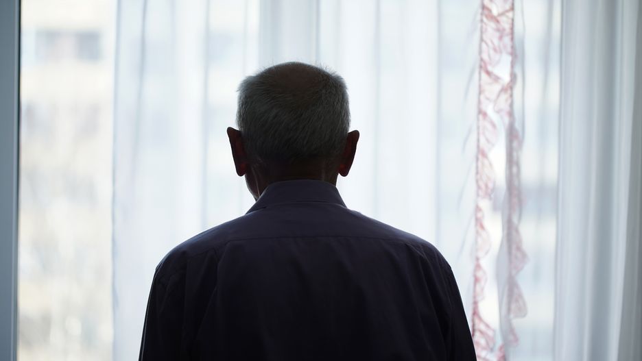Silhouette of retired man looking through window with transparent curtainSilhouette of retired man looking through window with transparent curtain standing at home rear view. Loneliness and old human care concept.Yaraslau Saulevich