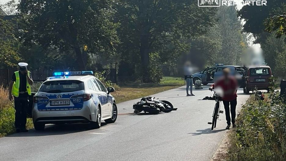 Policjant w kamizelce odblaskowej rozmawia przez telefon obok przewróconego motocykla na drodze, a rowerzysta prowadzi rower.